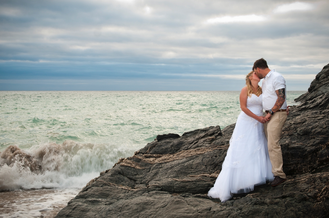 Wedded couple kiss in front of a stormy sky as waves crash onto the rocks