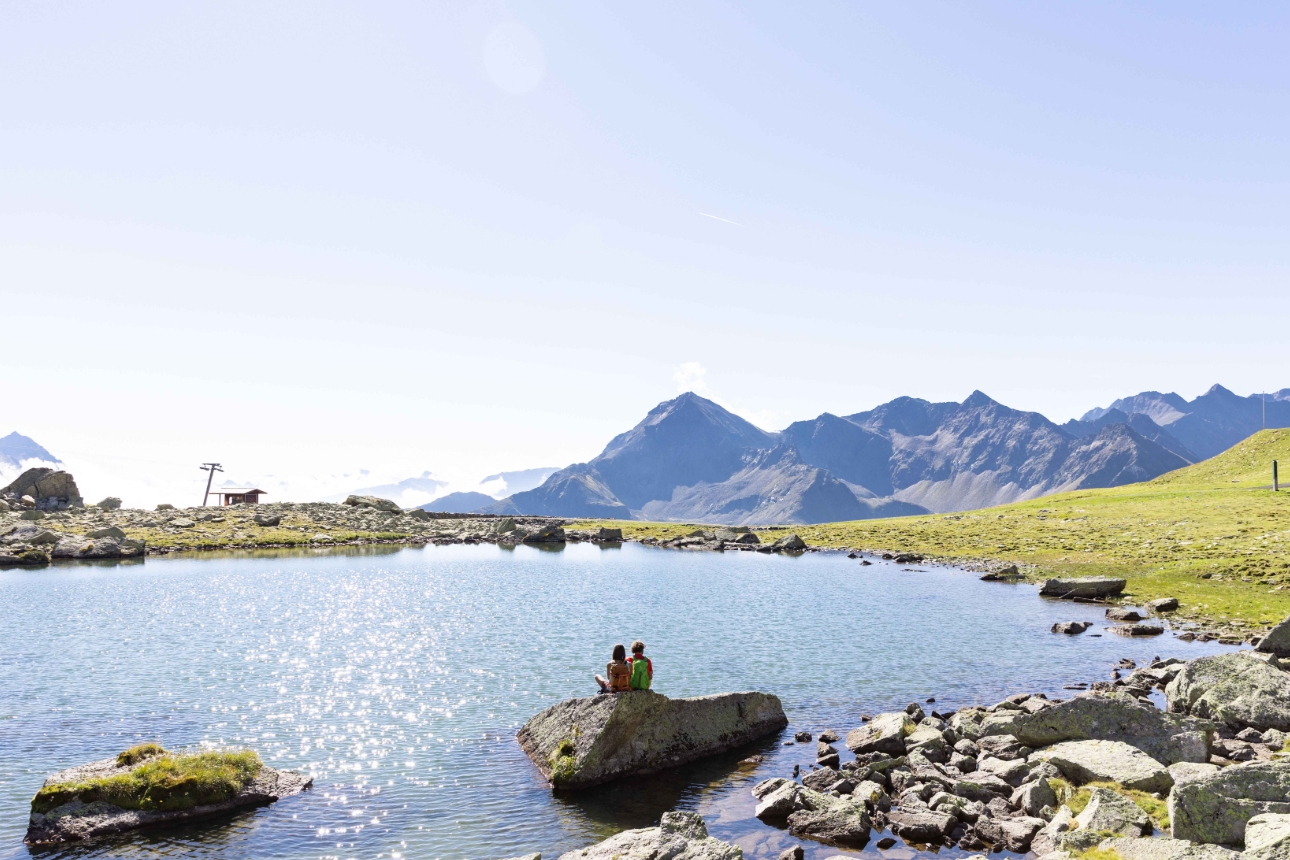 a lake looking out at mountains with two people sat on a very large rock